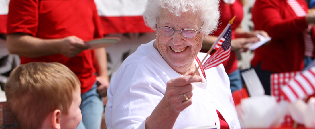 photo of senior woman waving an American flag at a 4th of July event and smiling at her grandson